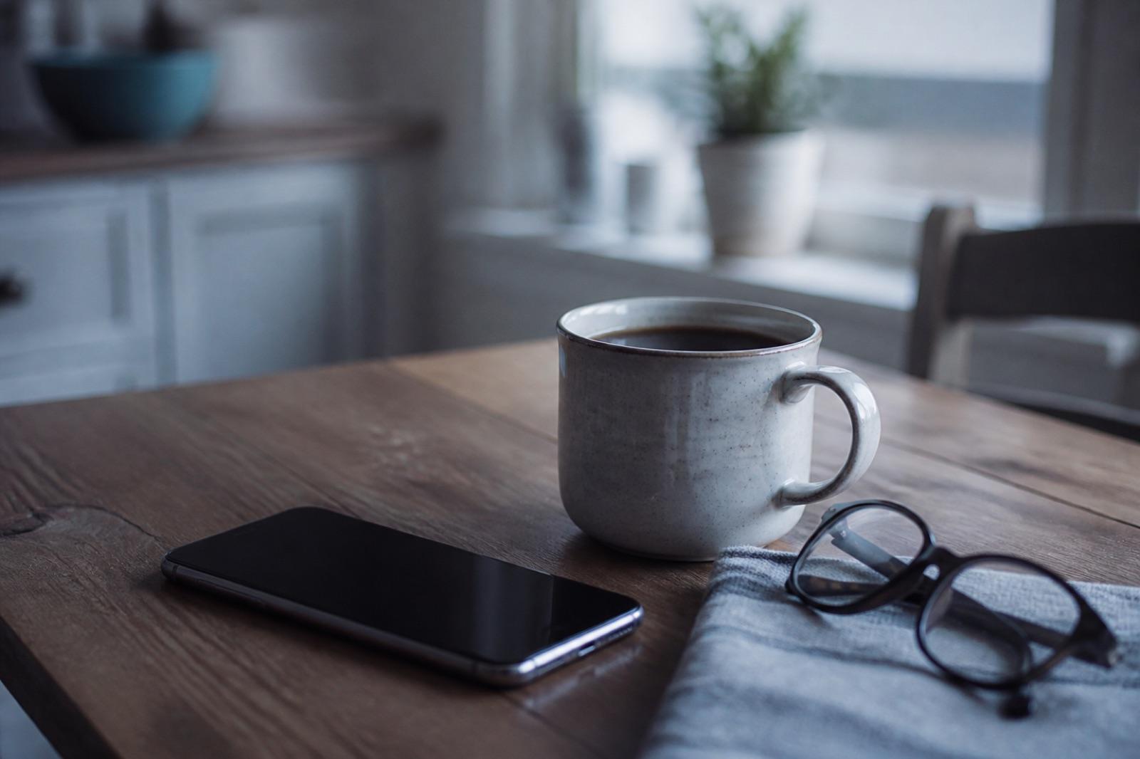 A kitchen table in the morning with a phone next to a cup of coffee